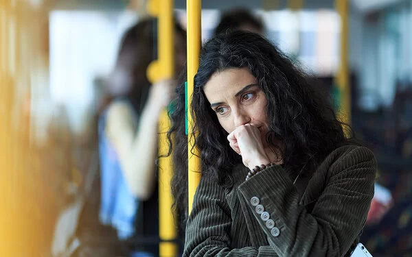 Eine junge Frau steht in einem Bus. Sie lehnt sich an eine gelbe Haltestange, dabei scheint sie intensiv zu grübeln.