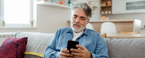 Mann mittleren Alters sitzt auf einem Sofa und schaut auf sein Smartphone.