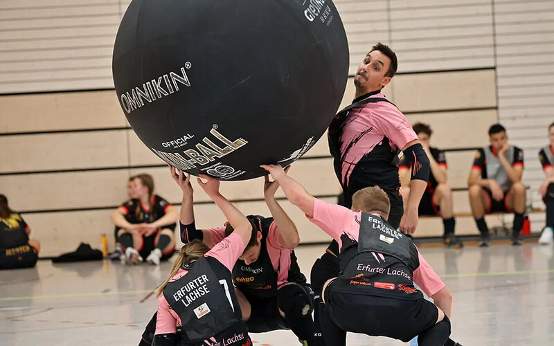 Drei Personen halten einen Kin-Ball in die Höhe, während ein Teammitglied den Ball mit der Brust anstößt.