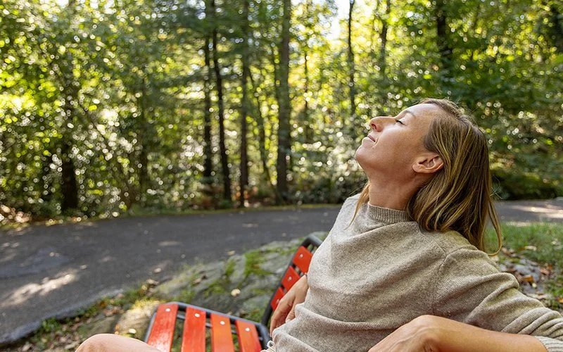 Auf einer Bank an einem von Bäumen gesäumten Weg sitzt eine Frau. Ihre Unterarme ruhen auf der Rückenlehne. Sie legt den Kopf in den Nacken und hat die Augen geschlossen.
