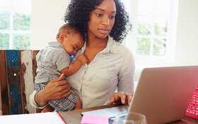 Junge Mutter am PC mit schlafendem Baby im Arm plant ihren beruflichen Wiedereinstieg nach der Elternzeit.