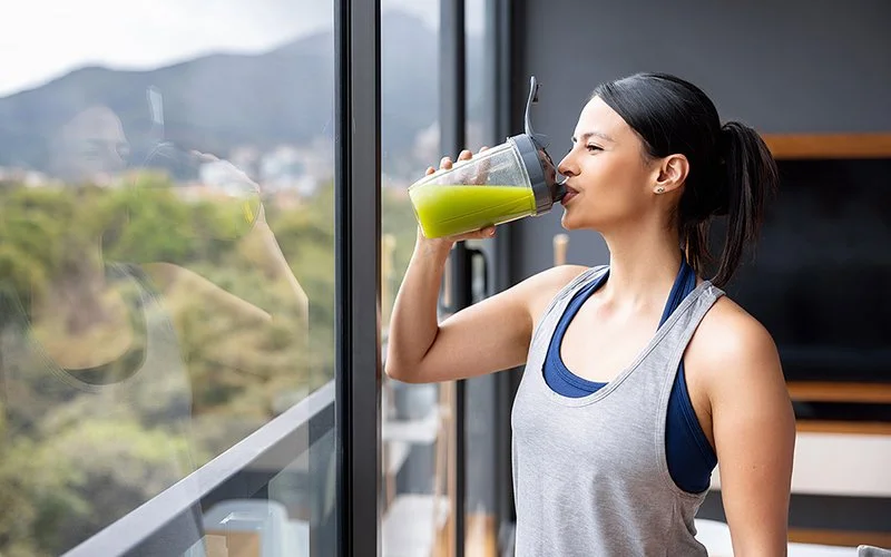 Eine sportliche Frau trinkt nach dem Training zu Hause einen grünen Gemüsesaft während sie aus einem großen Fenster blickt.