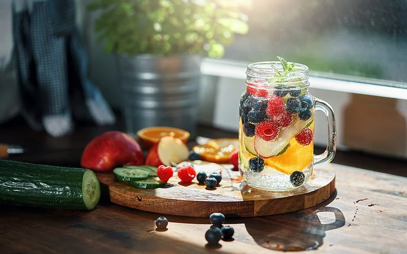 Auf einem runden Holzschneidebrett auf einer Küchenarbeitsplatte vor einem Fenster steht ein Henkelglas mit einem alkoholfreien Cocktail. Im Glas schwimmen Beeren, Obst- und Gurkenscheiben sowie ein Minzzweig. Auf dem Schneidebrett oder daneben liegen außerdem eine ganze Gurke, weitere Gurkenscheiben, Orangen, Apfelstücke, Himbeeren und Blaubeeren.