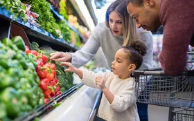 Ein Mädchen im Vorschulalter hilft ihren Eltern, Gemüse in der Obst- und Gemüseabteilung des Lebensmittelgeschäfts auszuwählen. Sie greift nach einer roten Paprika.