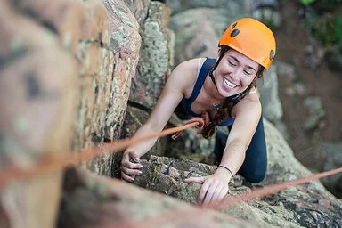 Eine junge Frau mit orangefarbenem Kletterhelm und Sicherheitsseil klettert lächelnd an einem felsigen Abhang empor.
