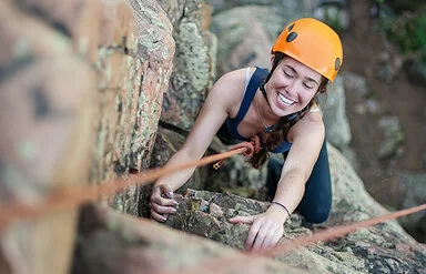 Eine junge Frau mit orangefarbenem Kletterhelm und Sicherheitsseil klettert lächelnd an einem felsigen Abhang empor.