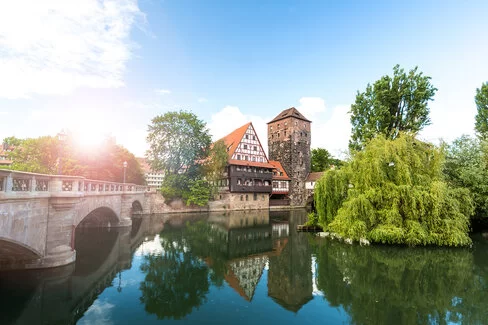 Das Bild zeigt zwei Häuser, die sich in einem See spiegeln. Rechts neben den Häusern steht eine Brücke. Auch sieht man ein paar große Bäume. Die Sonne scheint und der Himmel ist blau.