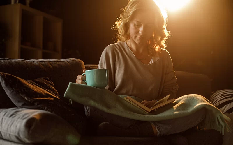 Eine Frau sitzt vor dem Schlafengehen bei gedimmtem Licht mit einem Buch auf dem Sofa.