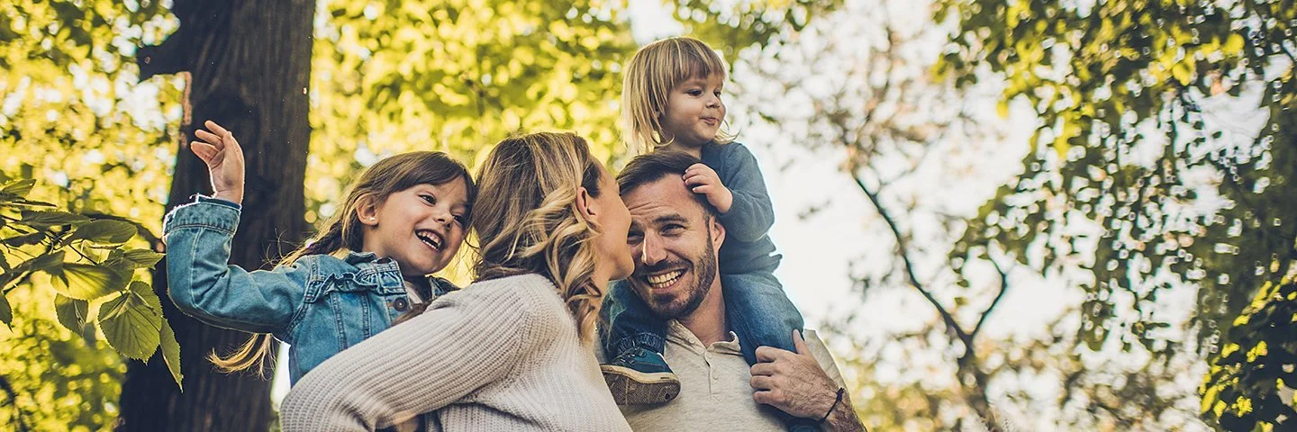 Eine junge vierköpfige Familie genießt unbeschwert einen Spaziergang im Wald. Die Eltern tragen die Kinder auf den Schultern.