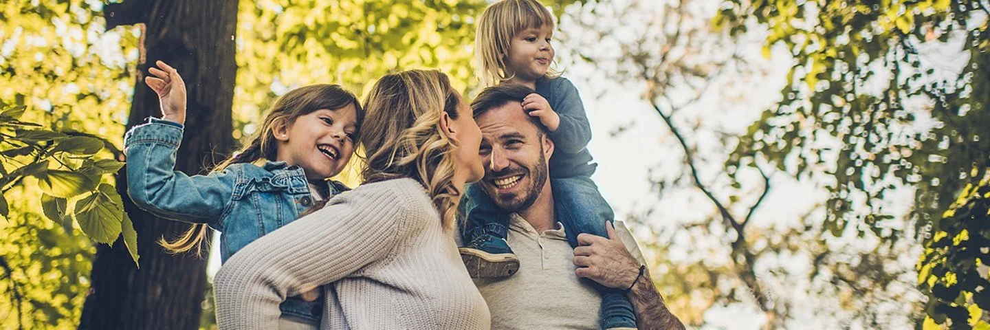 Eine junge vierköpfige Familie genießt unbeschwert einen Spaziergang im Wald. Die Eltern tragen die Kinder auf den Schultern.