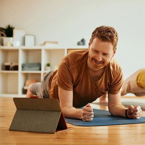 Ein Mann und eine Frau machen zu Hause auf Trainingsmatten Planks (Unterarmstütz-Übungen). Der Mann im Vordergrund betrachtet lächelnd das Display eines Tablets, das vor ihm aufgestellt ist.