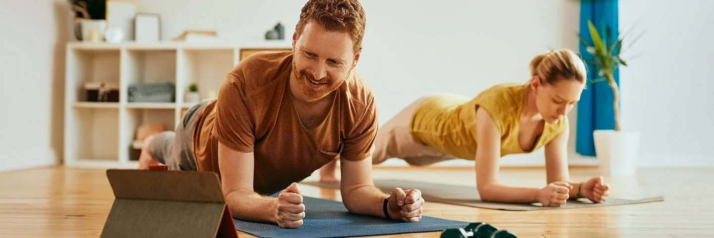 Ein Mann und eine Frau machen zu Hause auf Trainingsmatten Planks (Unterarmstütz-Übungen). Der Mann im Vordergrund betrachtet lächelnd das Display eines Tablets, das vor ihm aufgestellt ist.