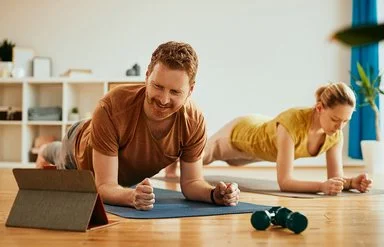 Ein Mann und eine Frau machen zu Hause auf Trainingsmatten Planks (Unterarmstütz-Übungen). Der Mann im Vordergrund betrachtet lächelnd das Display eines Tablets, das vor ihm aufgestellt ist.