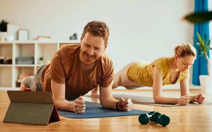 Ein Mann und eine Frau machen zu Hause auf Trainingsmatten Planks (Unterarmstütz-Übungen). Der Mann im Vordergrund betrachtet lächelnd das Display eines Tablets, das vor ihm aufgestellt ist.