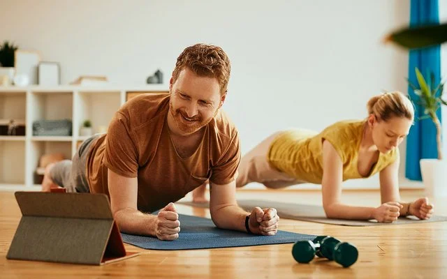 Ein Mann und eine Frau machen zu Hause auf Trainingsmatten Planks (Unterarmstütz-Übungen). Der Mann im Vordergrund betrachtet lächelnd das Display eines Tablets, das vor ihm aufgestellt ist.