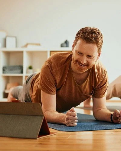 Ein Mann und eine Frau machen zu Hause auf Trainingsmatten Planks (Unterarmstütz-Übungen). Der Mann im Vordergrund betrachtet lächelnd das Display eines Tablets, das vor ihm aufgestellt ist.