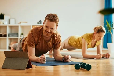 Ein Mann und eine Frau machen zu Hause auf Trainingsmatten Planks (Unterarmstütz-Übungen). Der Mann im Vordergrund betrachtet lächelnd das Display eines Tablets, das vor ihm aufgestellt ist.