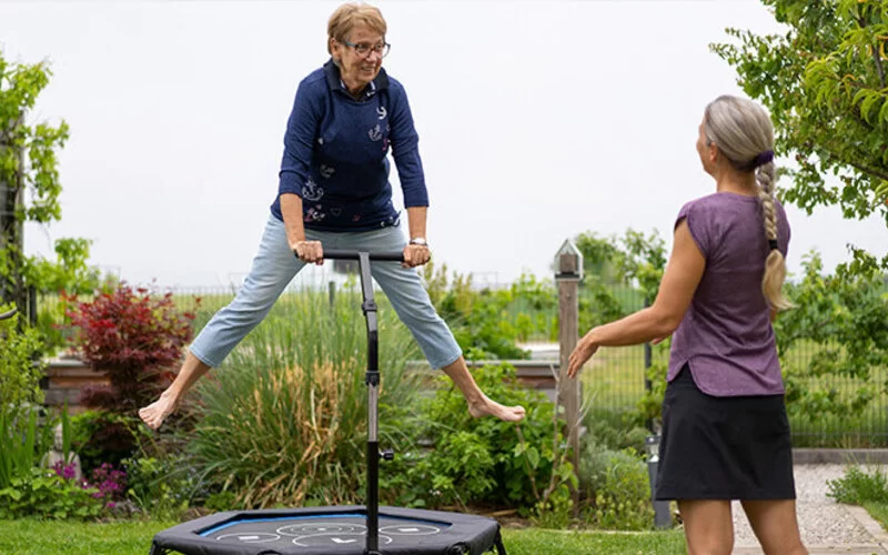 Ältere Frau macht Jumping Fitness auf einem Trampolin unter Anleitung einer Trainerin.