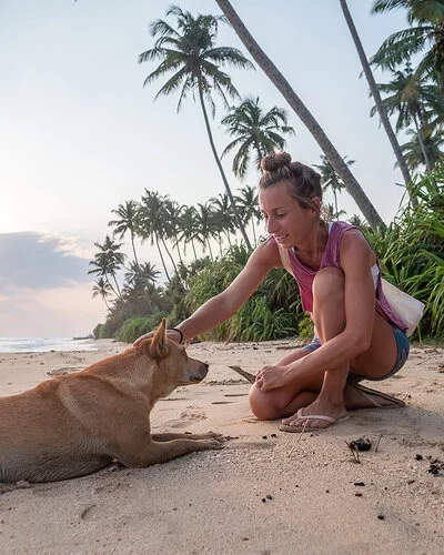 Eine blonde Frau streichelt an einem tropischen Strand einen Hund am Kopf.