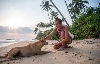 Eine blonde Frau streichelt an einem tropischen Strand einen Hund am Kopf.