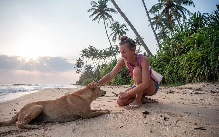 Eine blonde Frau streichelt an einem tropischen Strand einen Hund am Kopf.