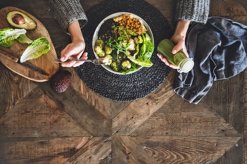 Ein Teller, gefüllt mit frischem Gemüse und Hülsenfrüchten, steht auf einem Holztisch. Vor dem Teller sitzt eine Person. Zu sehen sind nur die Hände. In der rechten Hand hält die Person eine Gabel, in der linken Hand eine kleine Flasche mit einem grünen Smoothie.