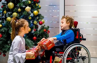Ein kleiner Junge im Rollstuhl packt gemeinsam mit seiner Schwester, die vor ihm hockt, Weihnachtsgeschenke vor einem geschmückten Weihnachtsbaum aus.