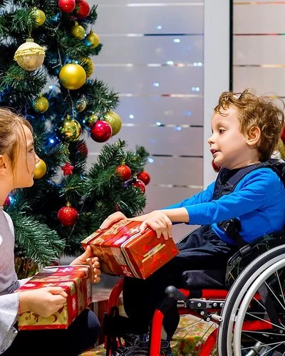 Ein kleiner Junge im Rollstuhl packt gemeinsam mit seiner Schwester, die vor ihm hockt, Weihnachtsgeschenke vor einem geschmückten Weihnachtsbaum aus.