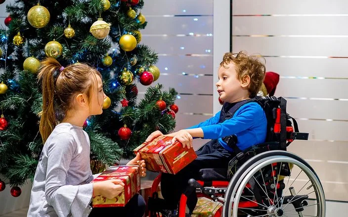 Ein kleiner Junge im Rollstuhl packt gemeinsam mit seiner Schwester, die vor ihm hockt, Weihnachtsgeschenke vor einem geschmückten Weihnachtsbaum aus.