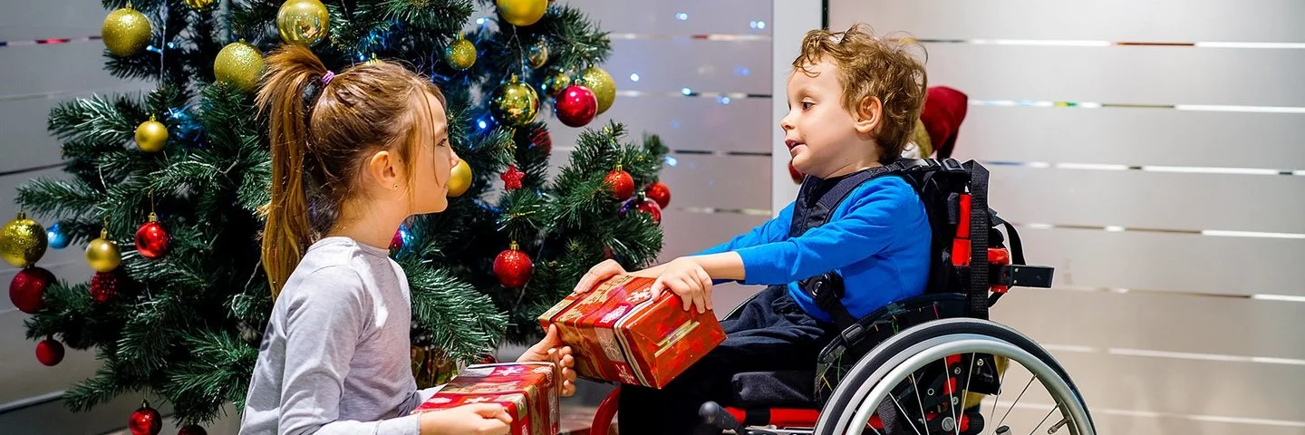 Ein kleiner Junge im Rollstuhl packt gemeinsam mit seiner Schwester, die vor ihm hockt, Weihnachtsgeschenke vor einem geschmückten Weihnachtsbaum aus.