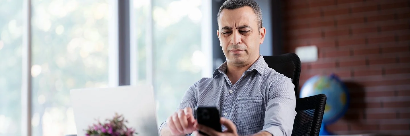Ein mittelalter Mann sitzt am Schreibtisch vor dem Laptop und versucht, etwas auf dem Handy zu lesen, indem er das Handy etwas weiter weg hält.