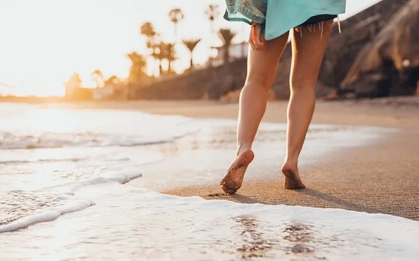 Eine Frau geht in Badekleidung barfuß an einem tropischen Sandstrand spazieren.