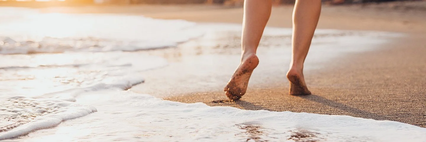 Eine Frau geht in Badekleidung barfuß an einem tropischen Sandstrand spazieren.