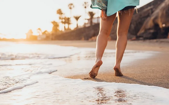 Eine Frau geht in Badekleidung barfuß an einem tropischen Sandstrand spazieren.