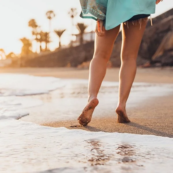 Eine Frau geht in Badekleidung barfuß an einem tropischen Sandstrand spazieren.