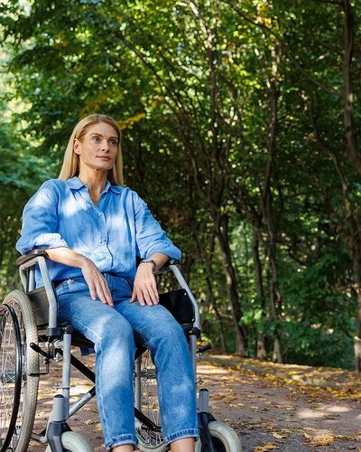 Eine Frau sitzt im Rollstuhl, im Hintergrund ein Wald.