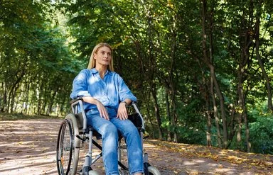 Eine Frau sitzt im Rollstuhl, im Hintergrund ein Wald.