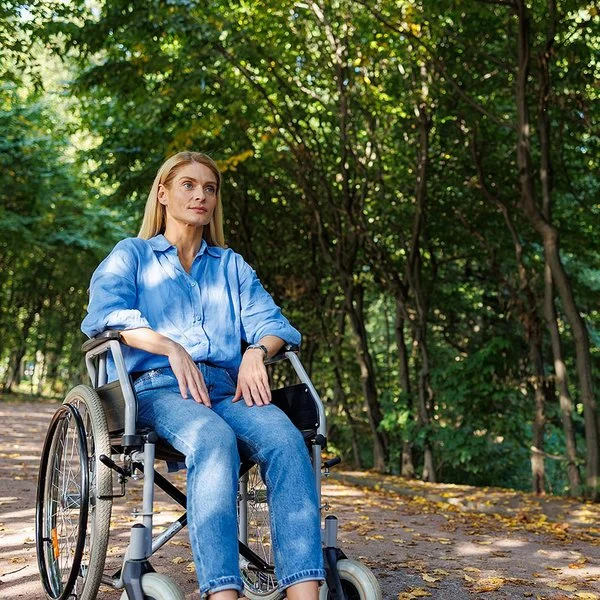 Eine Frau sitzt im Rollstuhl, im Hintergrund ein Wald.