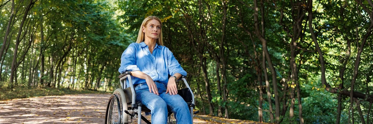 Eine Frau sitzt im Rollstuhl, im Hintergrund ein Wald.
