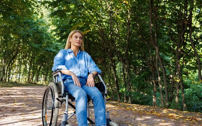 Eine Frau sitzt im Rollstuhl, im Hintergrund ein Wald.