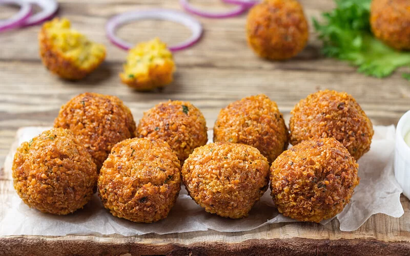 Falafelbällchen auf einem Holzbrettchen mit einer Dipschale, im Hintergrund liegen Zwiebelringe und Salat.