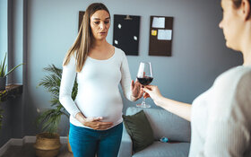 Eine schwangere Frau hält mit der rechten Hand ihren Babybauch, während sie ihre linke Hand vor ein Glas Wein hebt, das eine andere Frau ihr anbietet.