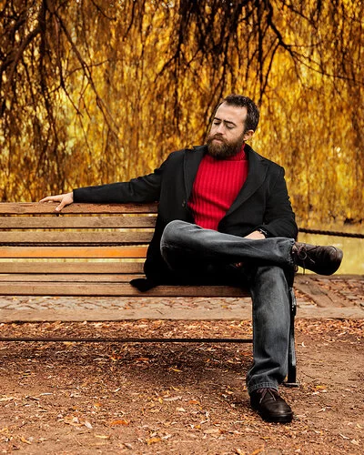 Ein Mann in einem roten Pullover und einer schwarzen Jacke sitzt alleine auf einer Parkbank.