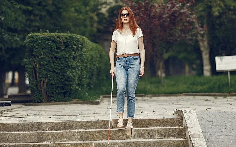 Eine junge Frau mit langen roten Haaren und Sonnenbrille geht mit einem Blindenstock eine Treppe in einem Park hinunter.