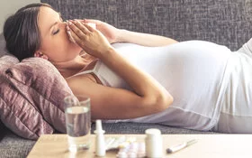 Eine schwangere Frau mit Erkältung liegt auf dem Sofa und putzt sich die Nase.
