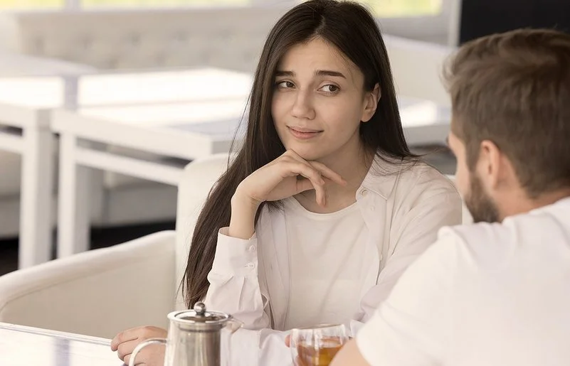 Eine Frau sitzt mit einem Mann (der nur von hinten zu sehen ist) in einem Café. Sie stützt ihr Kinn auf ihre Hand und blickt scheinbar gelangweilt, skeptisch und etwas genervt zu ihrem Gegenüber.