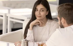 Eine Frau sitzt mit einem Mann (der nur von hinten zu sehen ist) in einem Café. Sie stützt ihr Kinn auf ihre Hand und blickt scheinbar gelangweilt, skeptisch und etwas genervt zu ihrem Gegenüber.