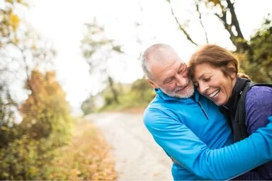 Ein Seniorenpaar ist draußen im Wald unterwegs. Sie lachen und umarmen sich.