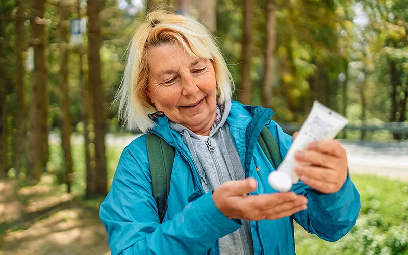 Frau in mittlerem Alter, mit Regenjacke und Rucksack in der Natur unterwegs, drückt aus einer Tube Creme in die Hand.