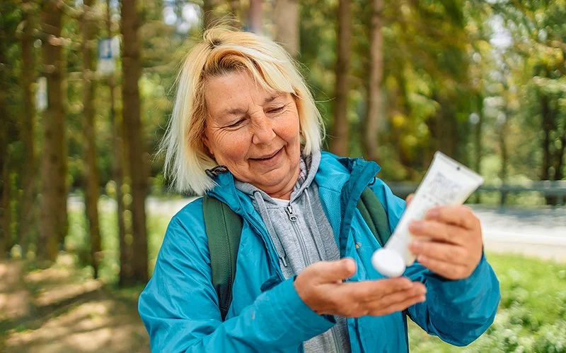 Frau in mittlerem Alter, mit Regenjacke und Rucksack in der Natur unterwegs, drückt aus einer Tube Creme in die Hand.